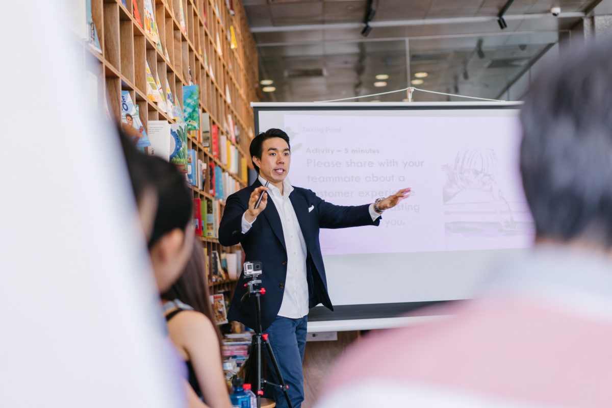 Image of a man standing next to a projector. He is teaching something to a group of people sitting in front of him