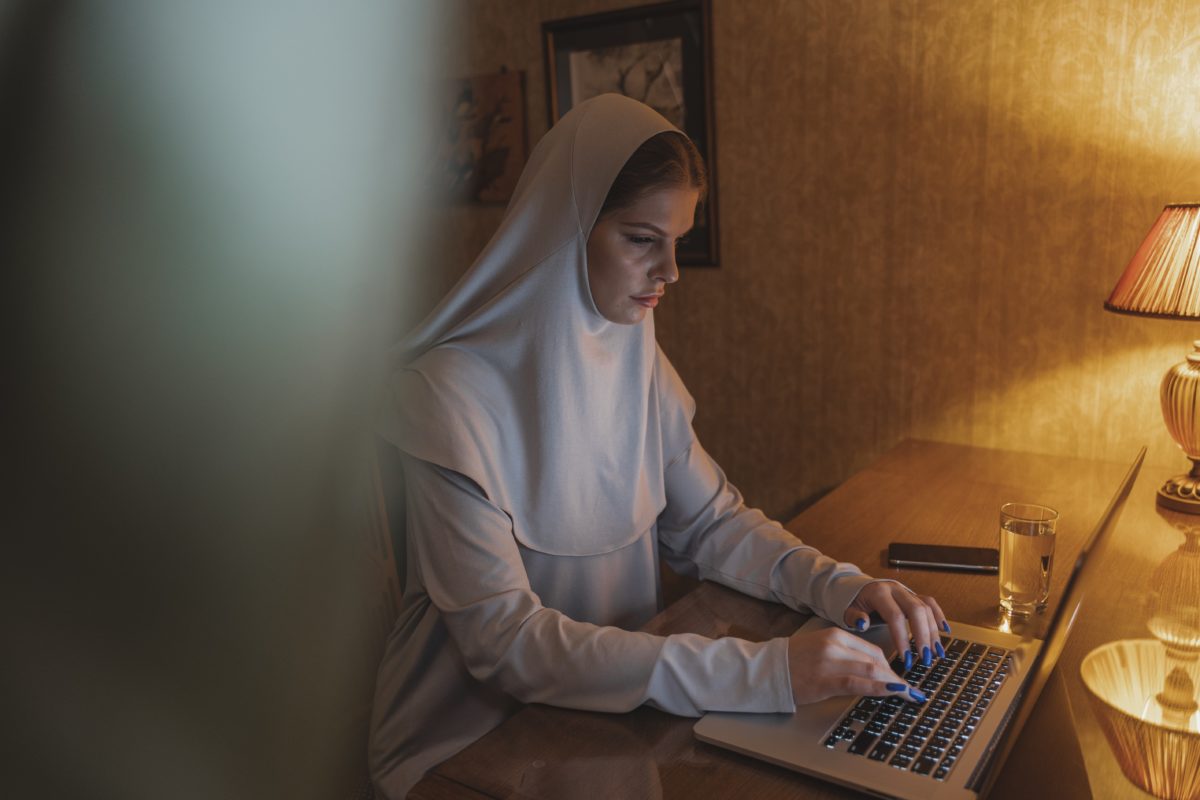 Image of woman in hijab sitting in a dark room on a laptop. There is a warm light coming off the lamp next to her.