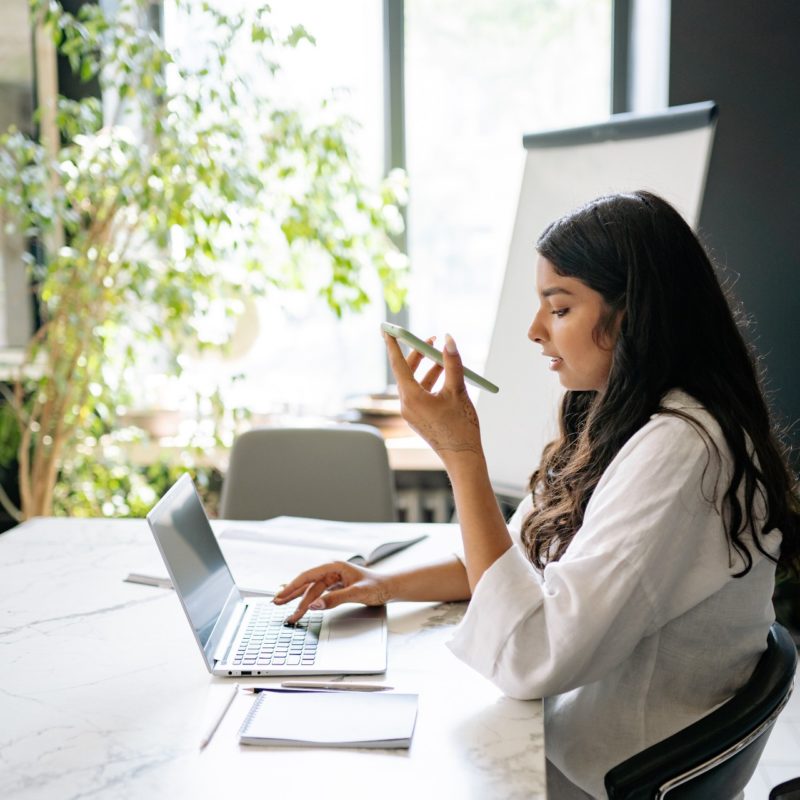 Image of a young woman sitting at her desk in front of a laptop taking a phone call. This is a body image for an article about unnecessary meetings.