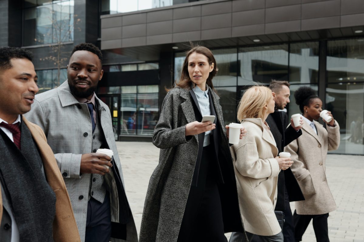 Image of six colleagues of mixed race walking in a line outside an office building. They're all holding coffee and talking to each other.