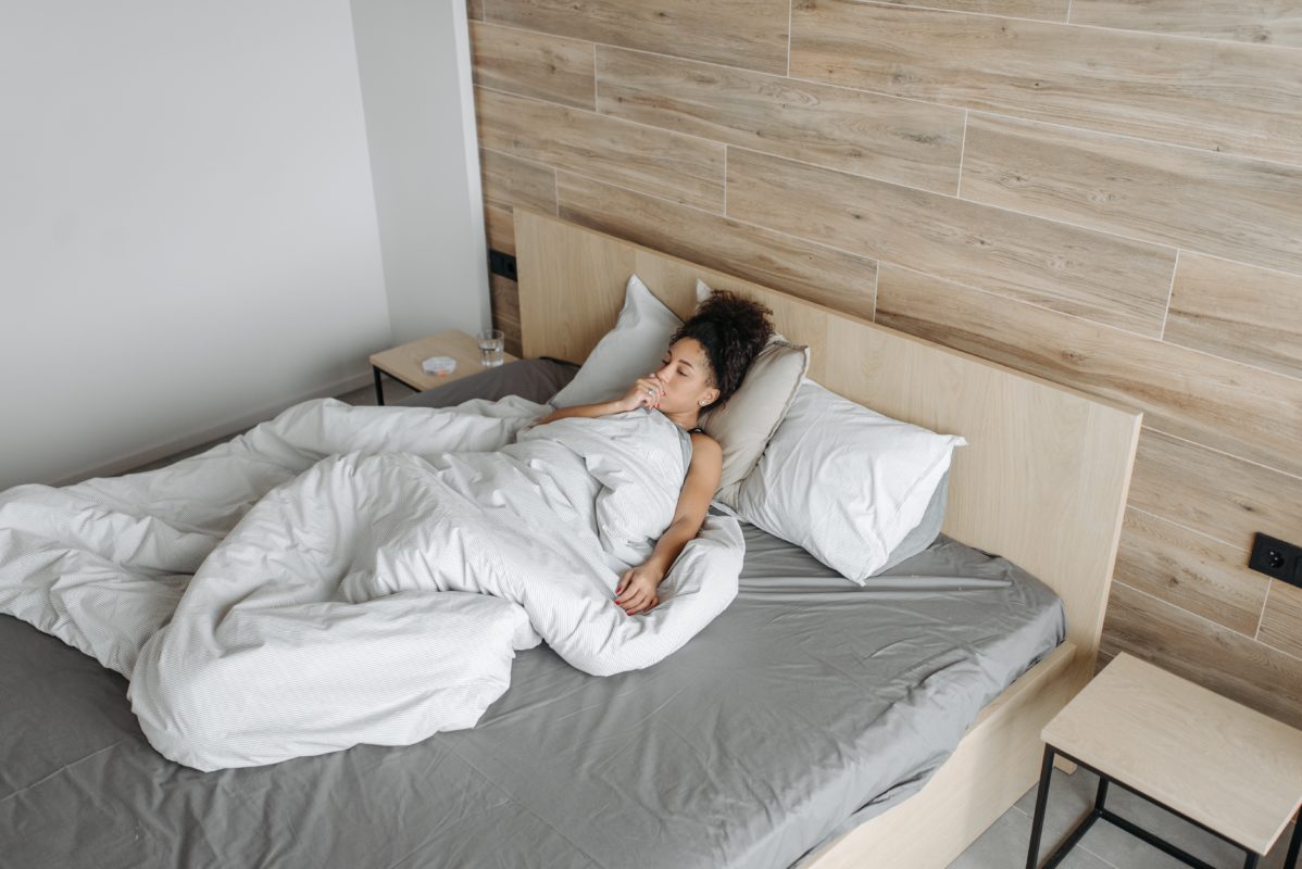 Woman lies under a doona in bed. She looks like she is coughing.