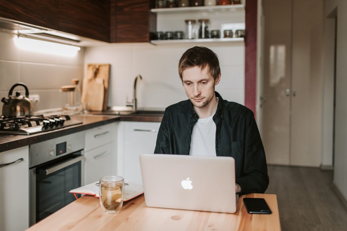 A young man sits at his kitchen counter on a laptop. His expression is neutral.