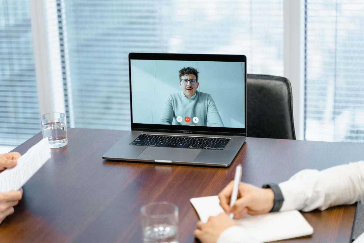 Image of a young man on a laptop screen sitting on a board room table. It looks like there are other people in the room