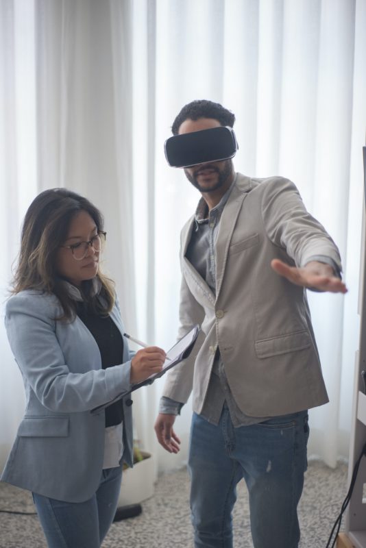 Photo of a man and woman. She is writing on a clipboard and he is wearing VR goggles.
